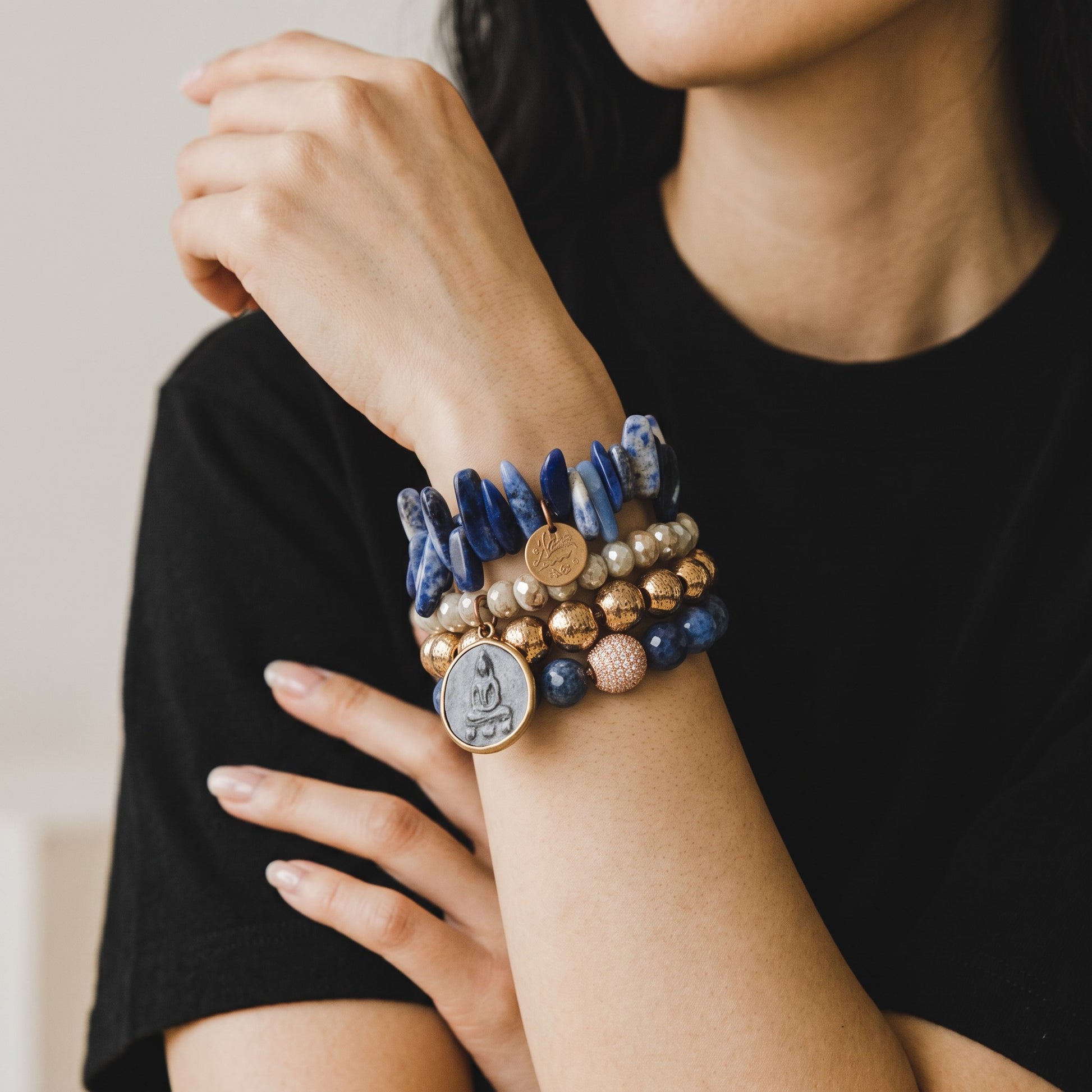 Trebor Style stackable bracelets featuring blue lapis, grey agate, and gold beads with a buddha charm on a wrist.